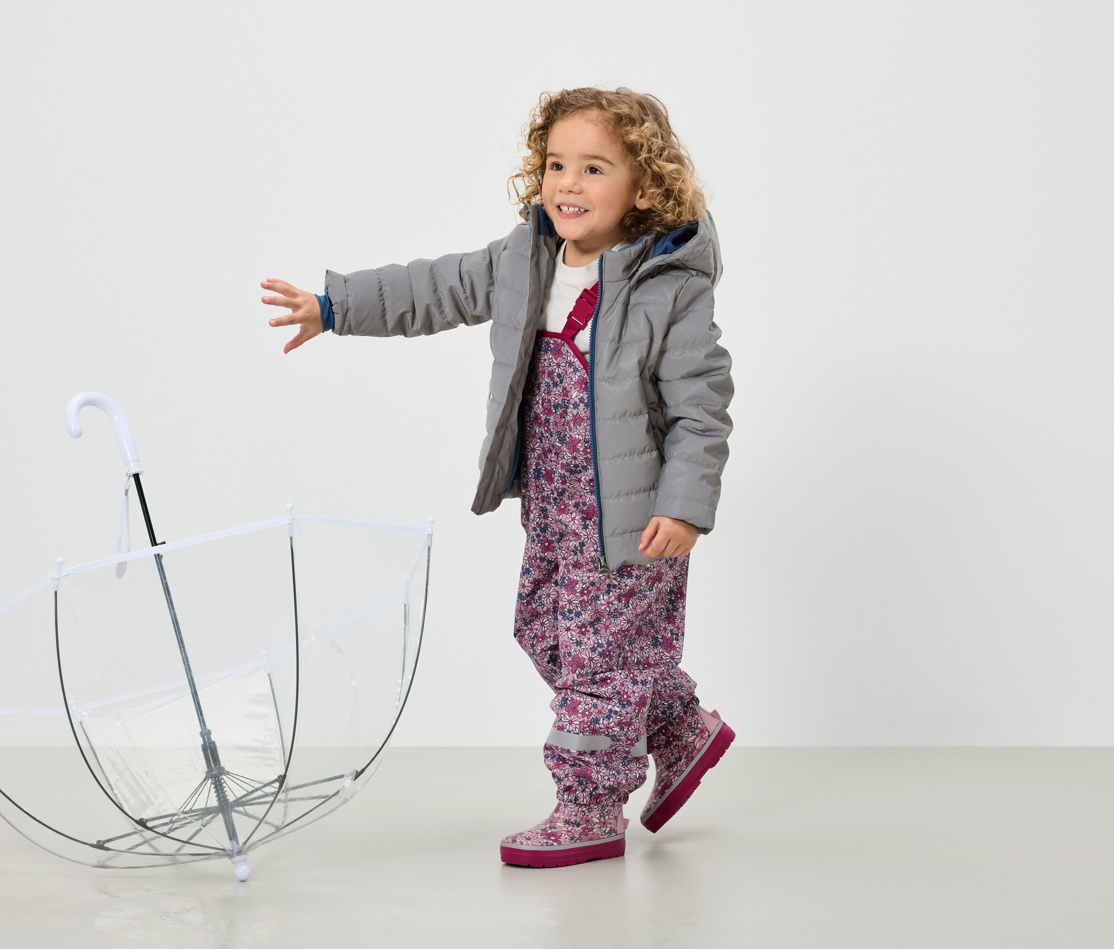 Uśmiechnięta, kręcona dziewczynka w szarej kurtce i kwiecistych spodniach stoi obok przezroczystego parasola.