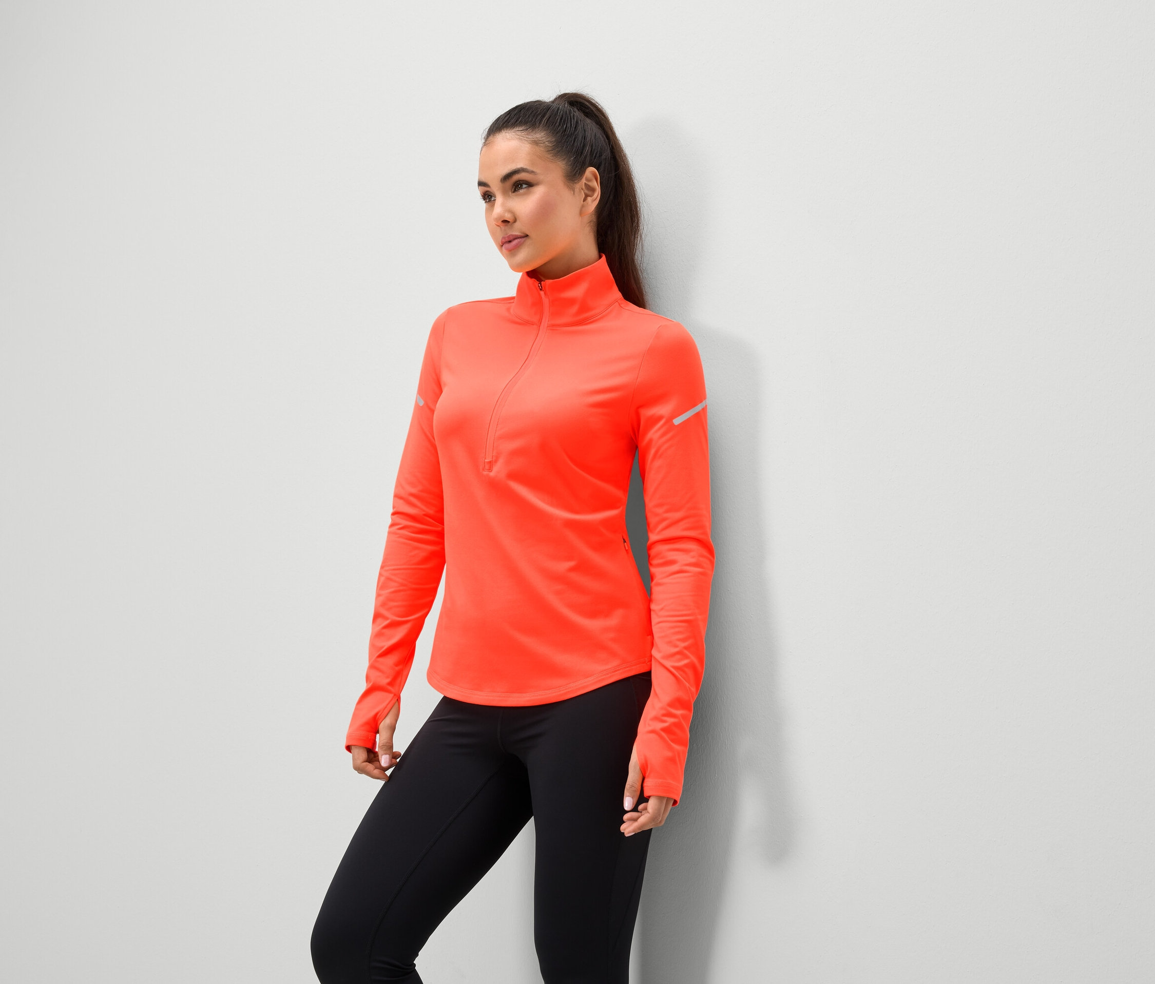 Kobieta w koralowej kurtce sportowej i czarnych legginsach stoi pod ścianą.