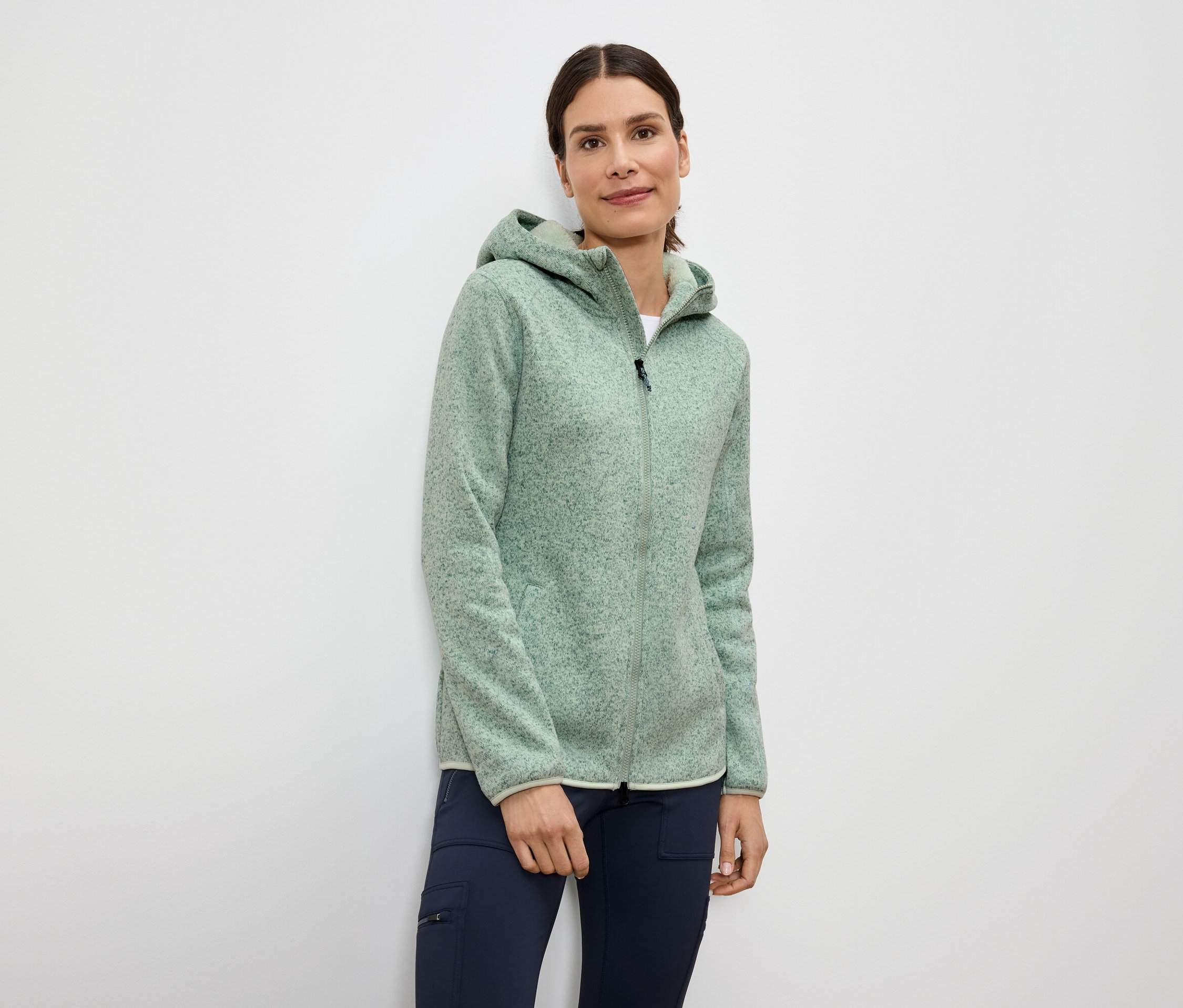 Kobieta ma na sobie jasnozieloną bluzę polarową z kapturem zapinaną na zamek i granatowe spodnie.