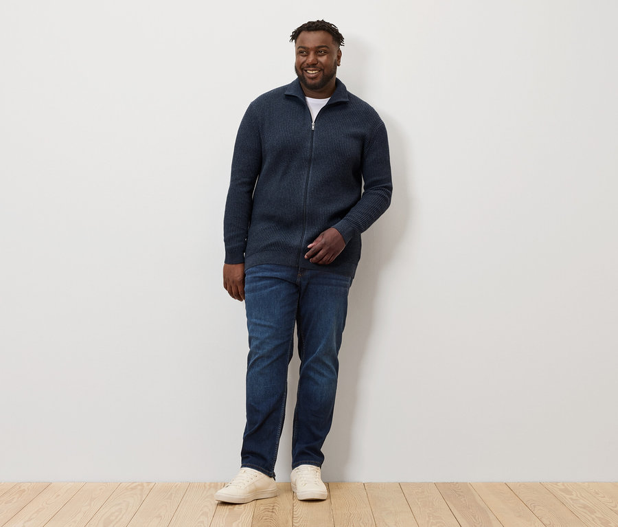 Mężczyzna stoi i ma na sobie rozpinany sweter z dzianiny, granatowy, dżinsy i tenisówki.