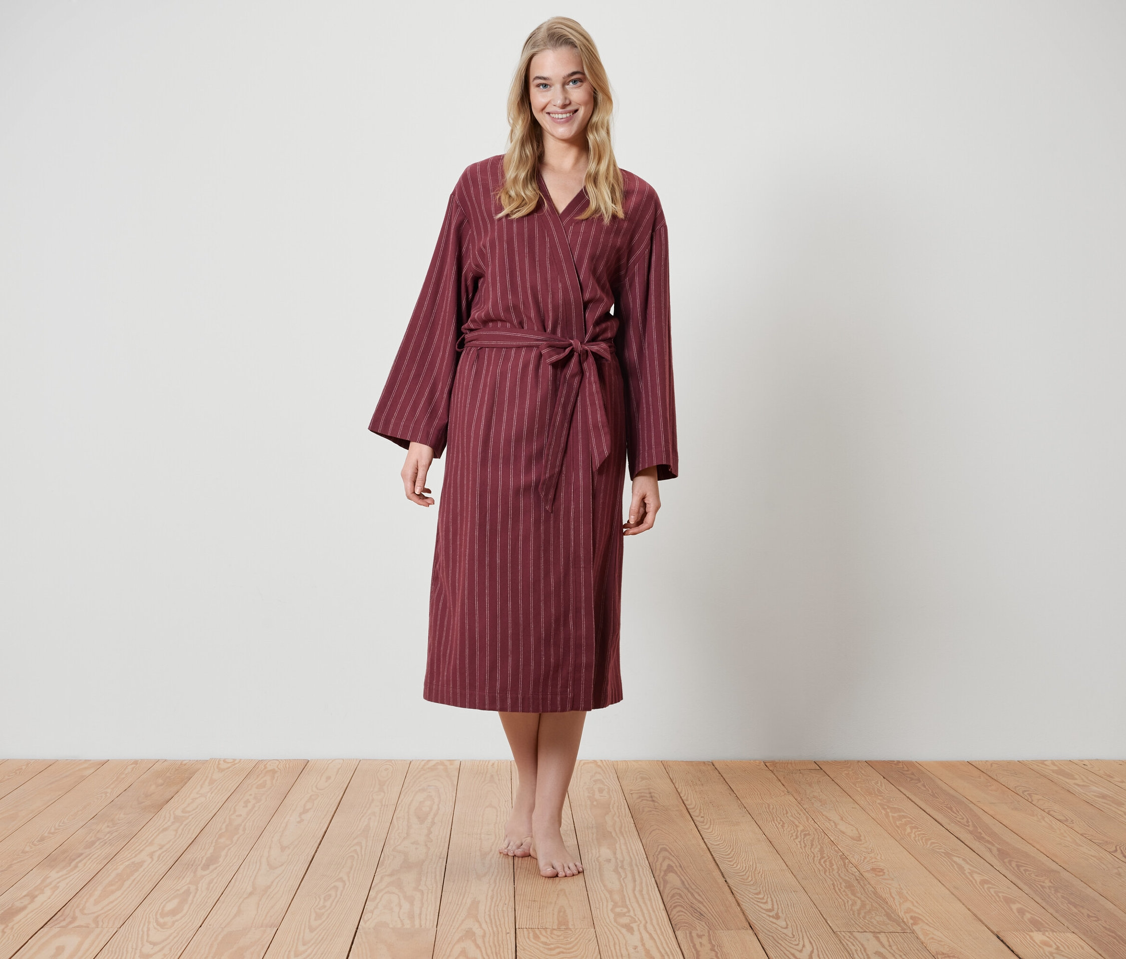 Blondynka stoi na drewnianej podłodze i ma na sobie bordowy szlafrok w paski.