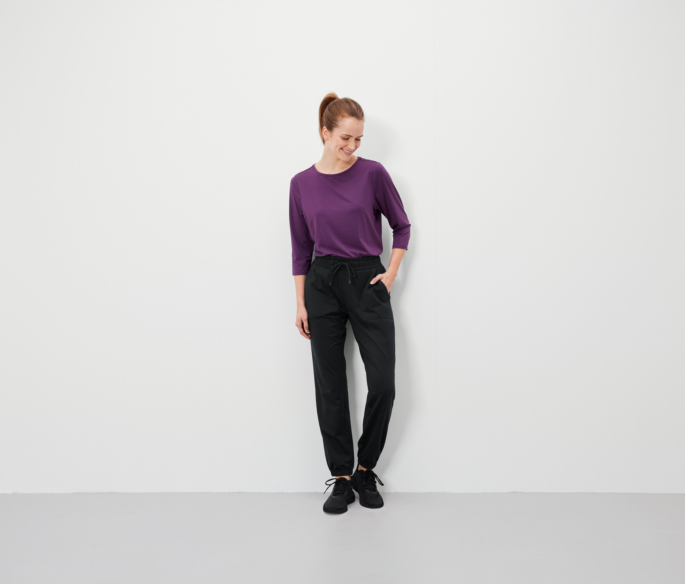 Kobieta z brązowymi włosami w kucyku stoi z rękami w kieszeniach czarnych spodni i ma na sobie fioletową koszulkę.