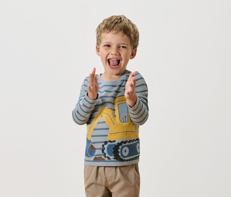 Chłopiec z blond włosami klaszcze w dłonie, ubrany w bluzkę z długim rękawem w niebieskie paski z żółtym buldożerem.