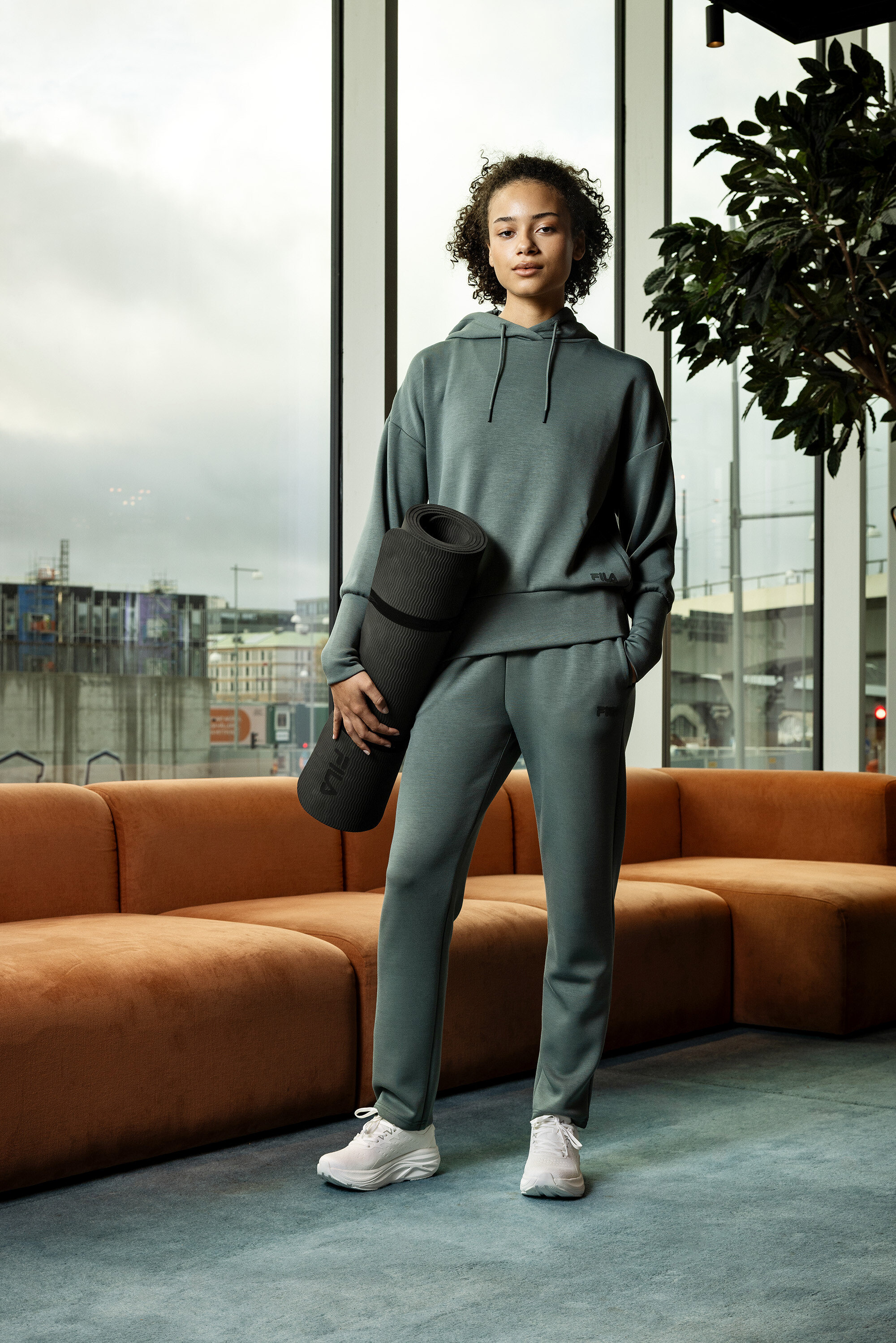 Młoda kobieta w szarym stroju sportowym trzyma zwiniętą matę do jogi na tle okna.