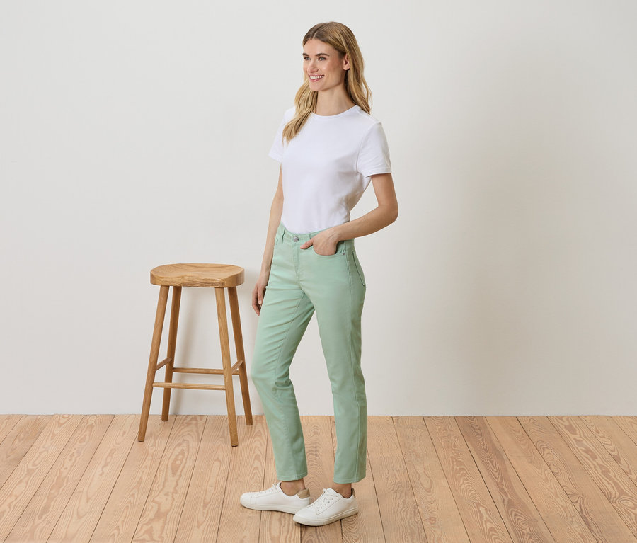 Kobieta stoi obok stołka, ma na sobie białą koszulkę, zielone spodnie i białe tenisówki.