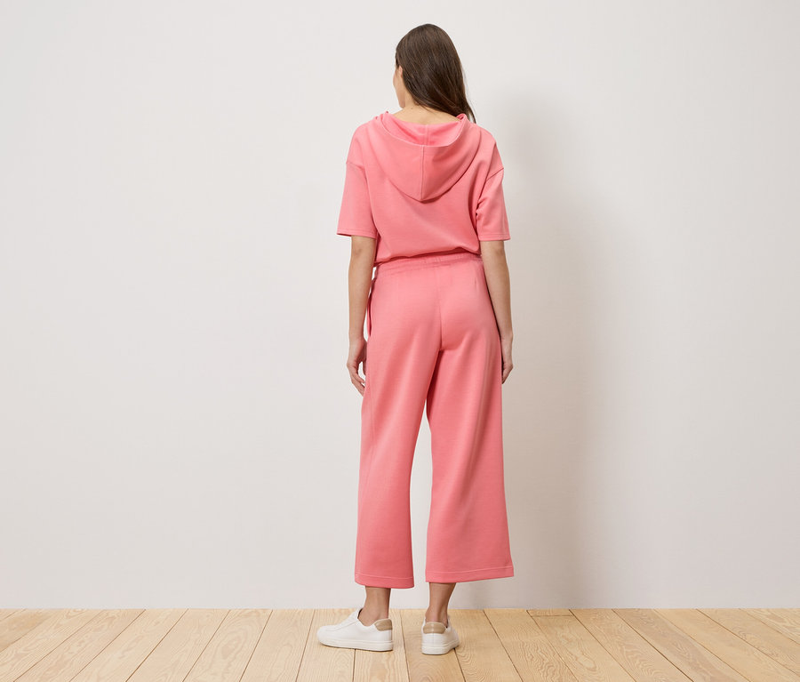 Kobieta stoi tyłem do kamery, ubrana w różową bluzę z kapturem i spodnie typu culottes.