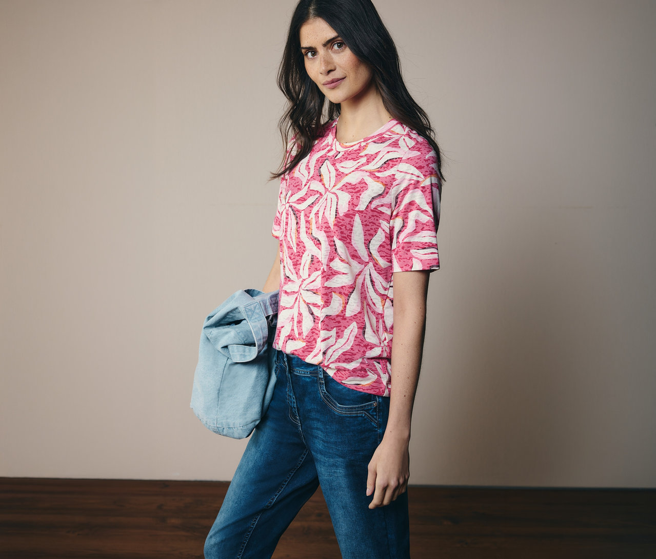 Kobieta ma na sobie różową koszulkę z krótkim rękawem CECIL we wzory i dżinsy. Na ramieniu ma kurtkę dżinsową.
