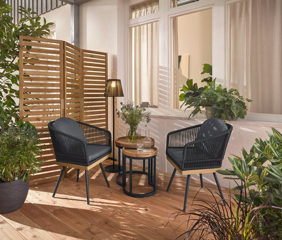 Dwa obrotowe krzesła ogrodowe z plecionką tekstylną stoją na drewnianym tarasie, pomiędzy nimi dwa stoliki ogrodowe z odłączanymi tacami, Ø ok. 40 cm i 50 cm. W tle elastyczny parawan i akumulatorowa lampa solarna.