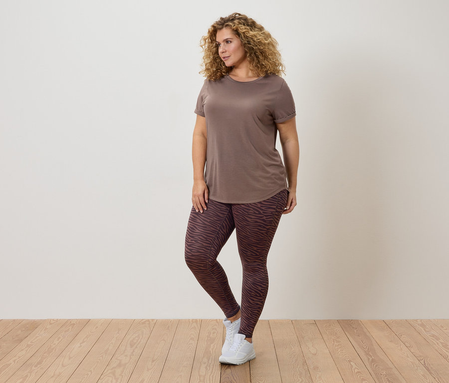 Kobieta z kręconymi włosami w brązowym t-shircie i wzorzystych legginsach pozuje na tle ściany.