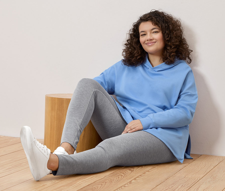 Kobieta o kręconych włosach siedzi na podłodze, ubrana w jasnoniebieską bluzę z kapturem, szare legginsy i białe tenisówki.