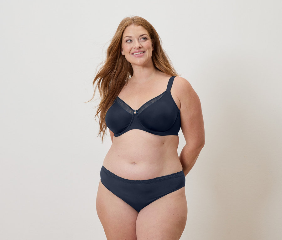 Kobieta z długimi rudymi włosami w granatowym biustonoszu i majtkach, uśmiecha się.