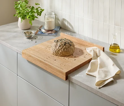 Na drewnianej desce na blacie kuchennym leży bochenek chleba z pestkami dyni. Obok chleba leży beżowa ściereczka.