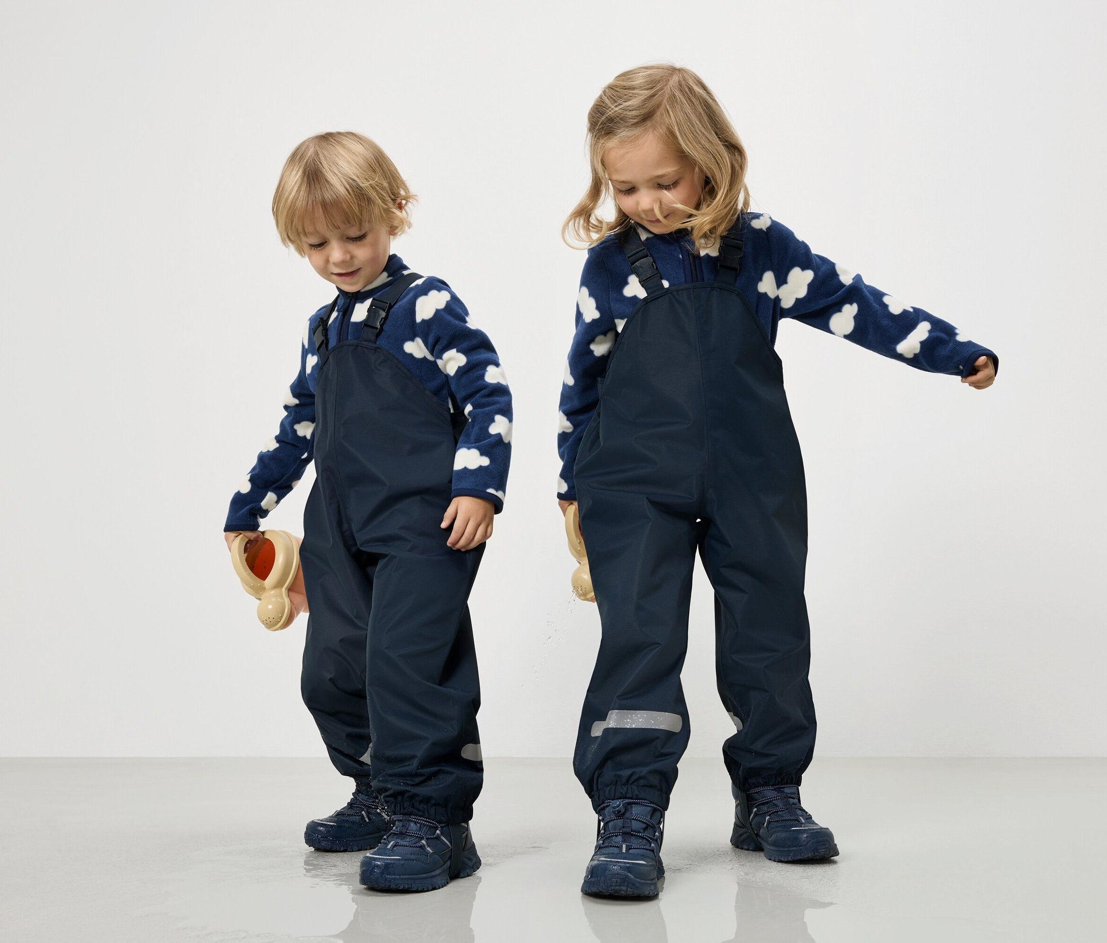 Dwoje dzieci w granatowych kombinezonach i niebiesko-białych bluzach w chmury stoi na białym tle.