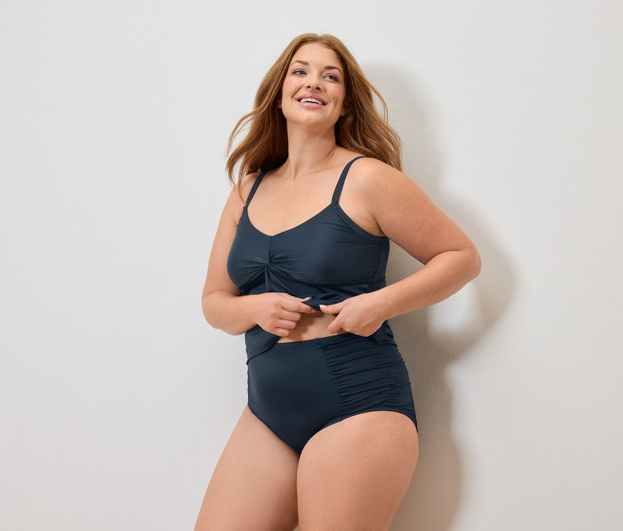 Kobieta z długimi rudymi włosami w granatowym kostiumie kąpielowym uśmiecha się.