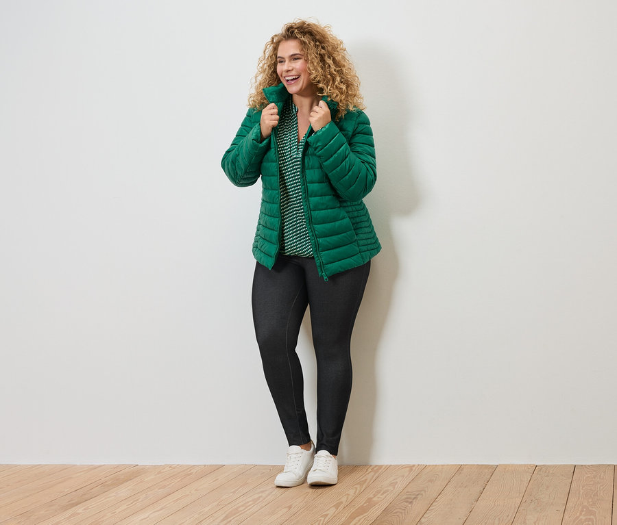 Uśmiechnięta kobieta z kręconymi włosami w zielonej kurtce, bluzce w paski, ciemnych legginsach i białych tenisówkach.