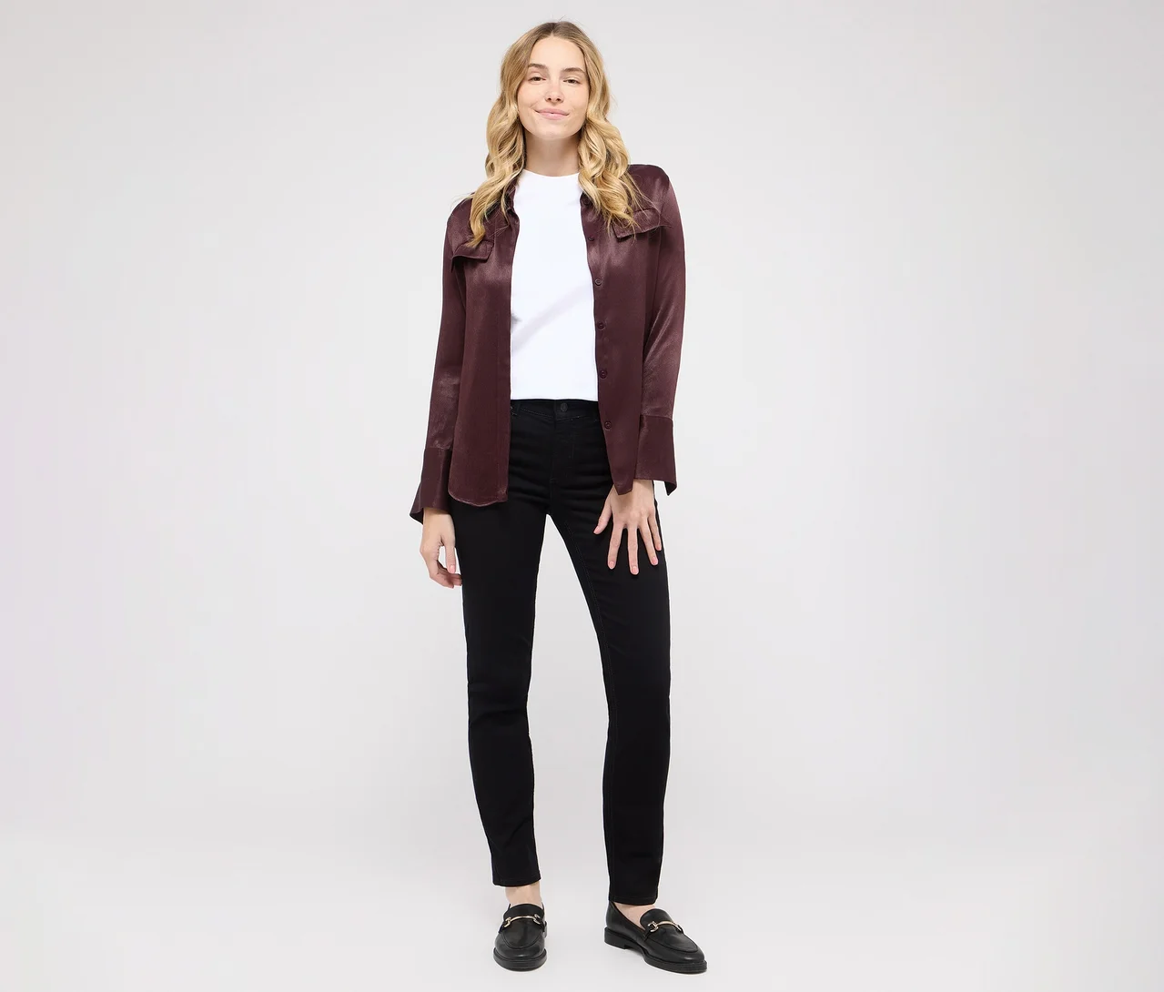 Kobieta z blond włosami stoi uśmiechnięta na białym tle. Ma na sobie biały T-shirt, rozpiętą bordową koszulę, czarne dżinsy skinny i czarne mokasyny ze złotymi klamrami.