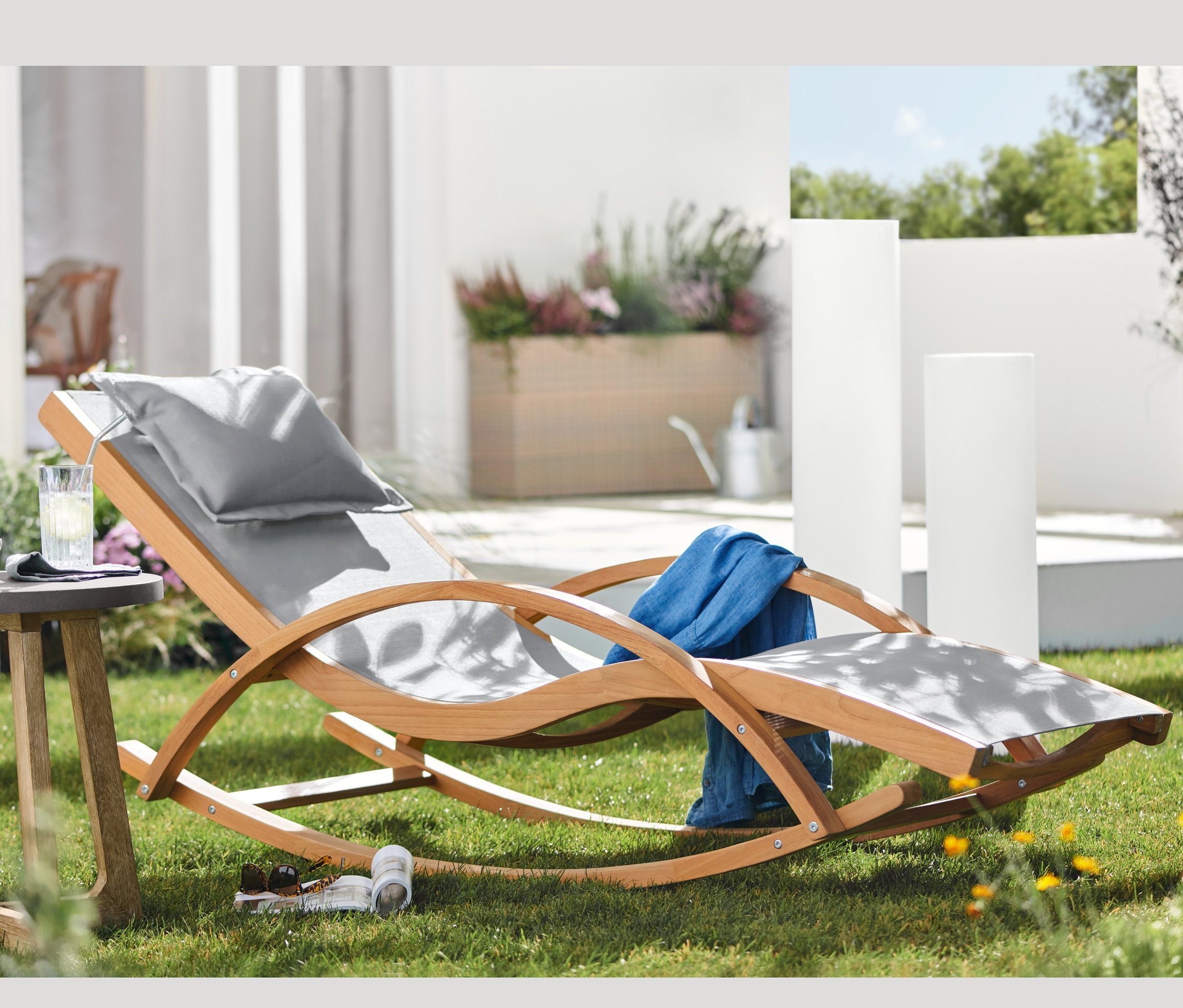 Na trawie stoi leżak na biegunach z drewna tekowego, z tkaniną, okrągły stolik z betonowym blatem i słupki solarne z przepływem kolorów o różnych wysokościach.