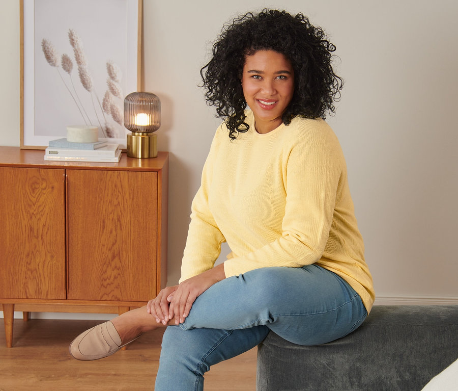 Kobieta siedzi na szarej sofie w żółtym swetrze z dzianiny, tregginsach i półbutach ze skóry welurowej.