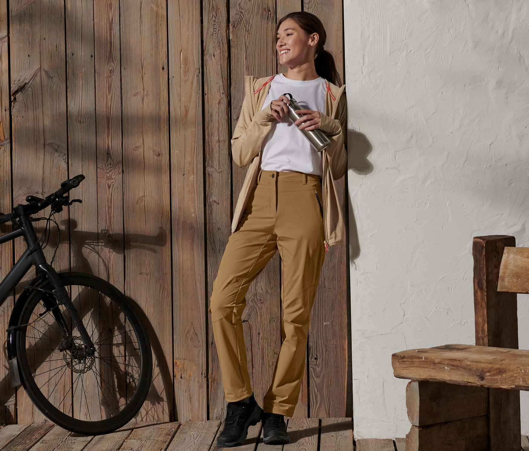 Uśmiechnięta kobieta trzyma butelkę wody ze stali nierdzewnej, opiera się o drewnianą ścianę i ma na sobie spodnie softshell.