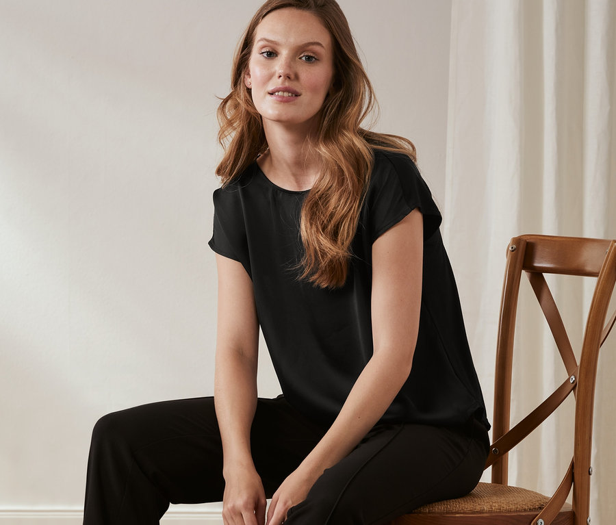 Kobieta siedzi na krześle, ubrana w czarne spodnie i bluzkę z satynowym przodem.