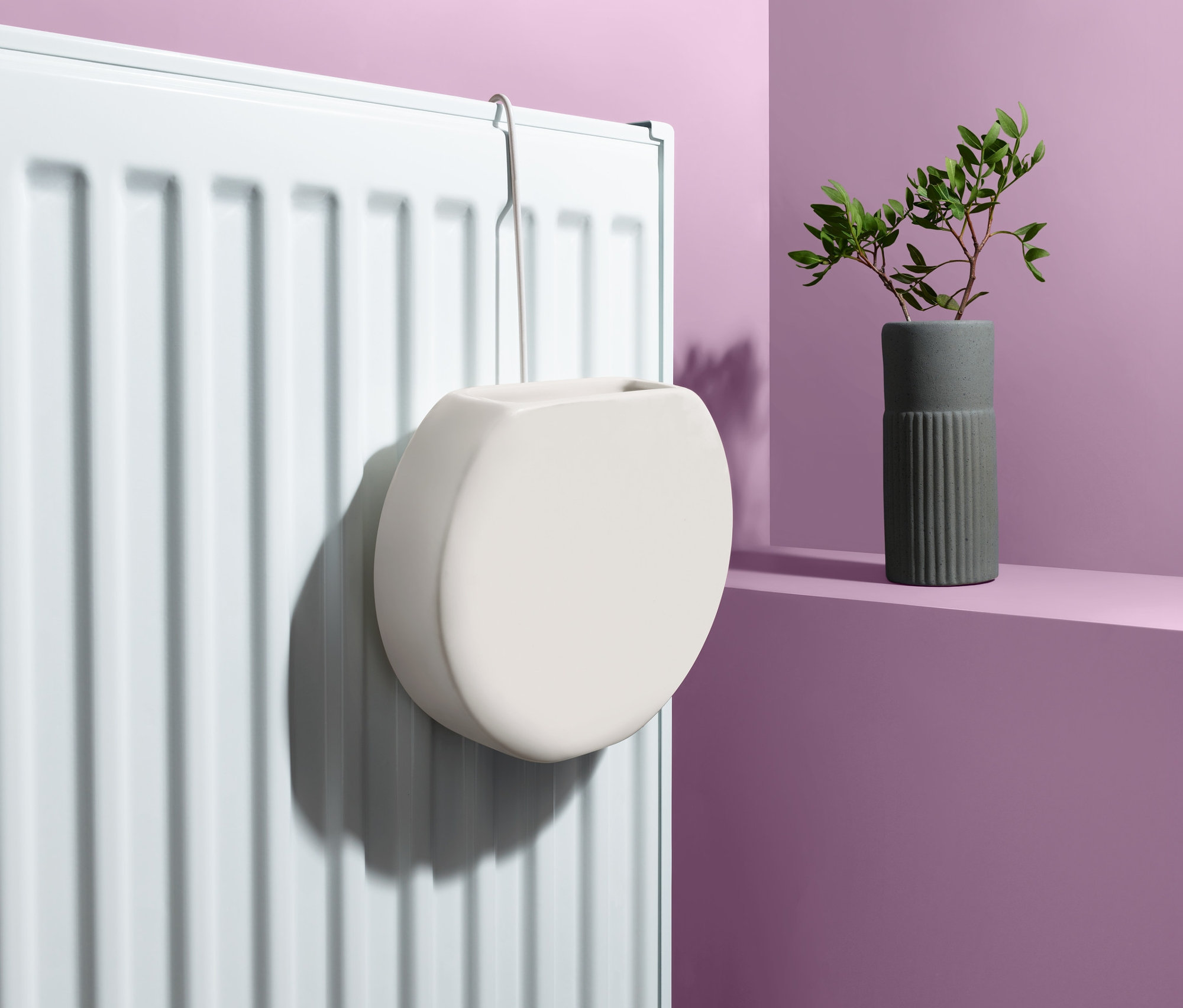 Biały nawilżacz powietrza na kaloryfer zawieszony na białym kaloryferze. Obok stoi wazon z zielonymi liśćmi.