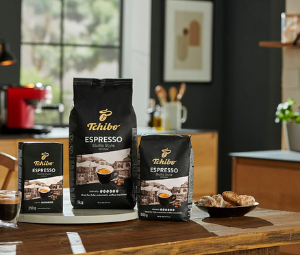 Trzy opakowania kawy Tchibo Espresso Sicilia Style (1 kg, 500 g, 250 g) stoją na drewnianym stole obok filiżanki espresso i talerza ciasteczek.