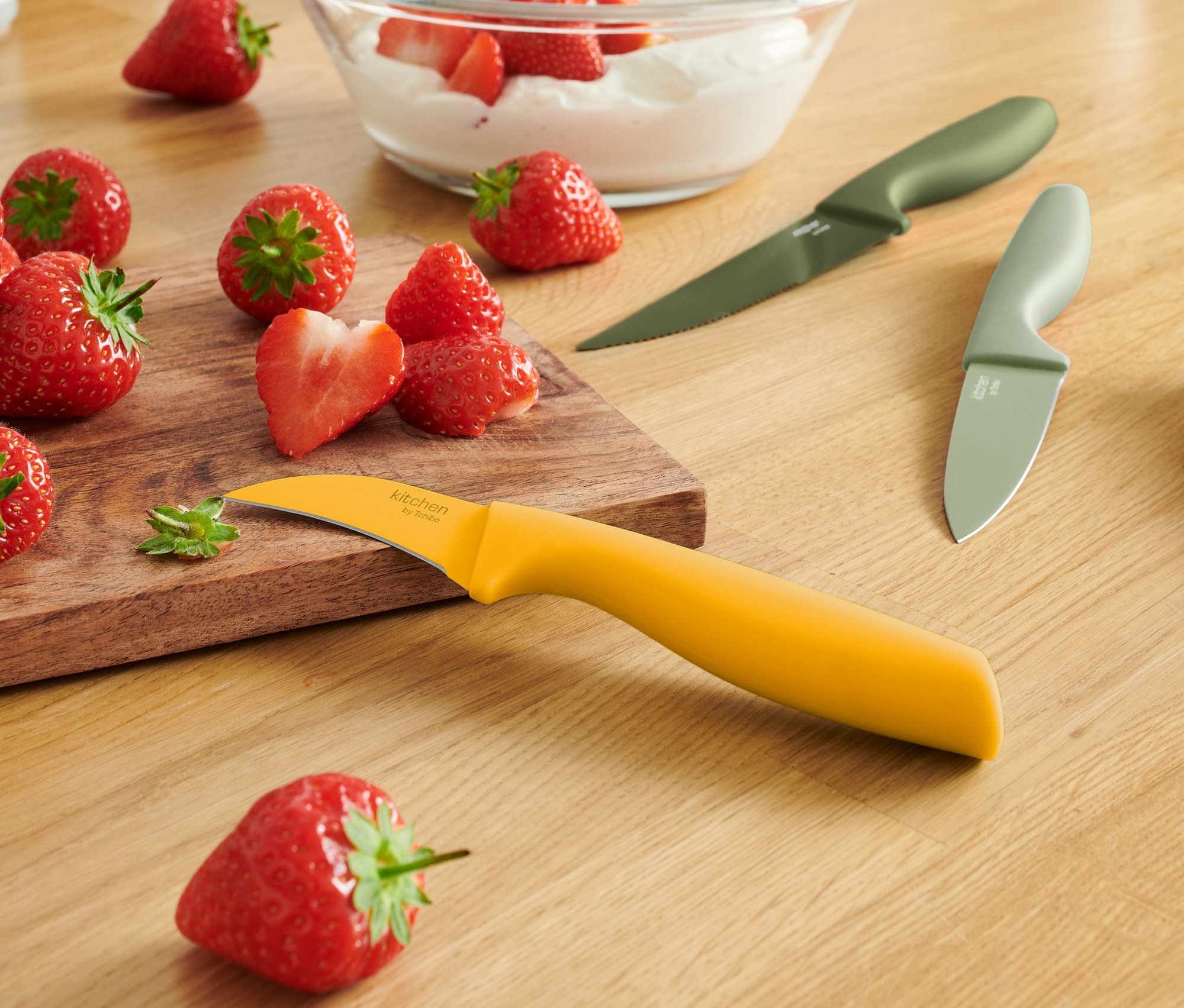 Na drewnianym stole leżą truskawki, miska z truskawkami i śmietaną oraz noże kuchenne, 3 sztuki.