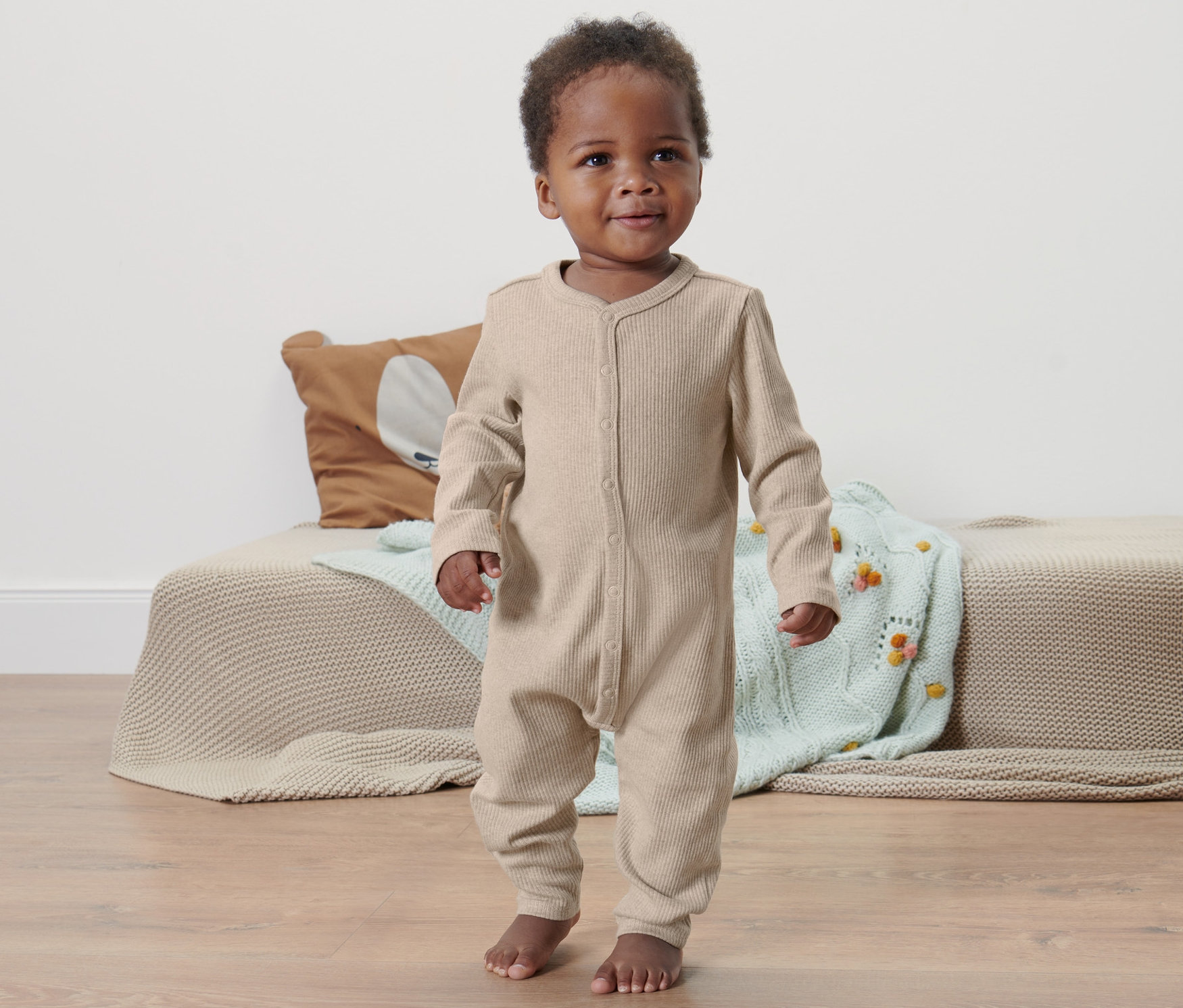 Dziecko o ciemnej karnacji stoi na podłodze i ma na sobie rampers niemowlęcy.