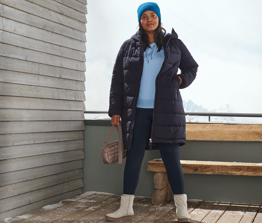 Kobieta pozuje na drewnianym tarasie w granatowym płaszczu pikowanym, niebieskiej czapce z dzianiny, bluzie dresowej z kapturem, niebieskich ocieplanych legginsach, botkach ze skóry zamszowej i z pikowaną torbą na ramię.