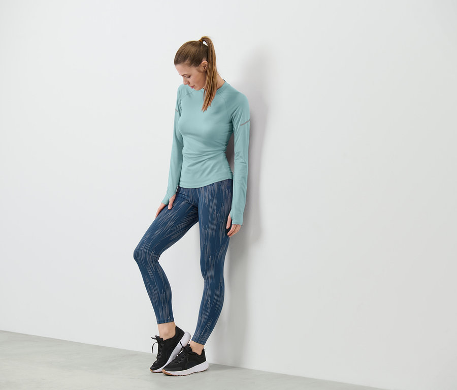 Kobieta oparta o ścianę ma na sobie legginsy sportowe ze srebrnym odblaskowym nadrukiem i bluzkę z długim rękawem.