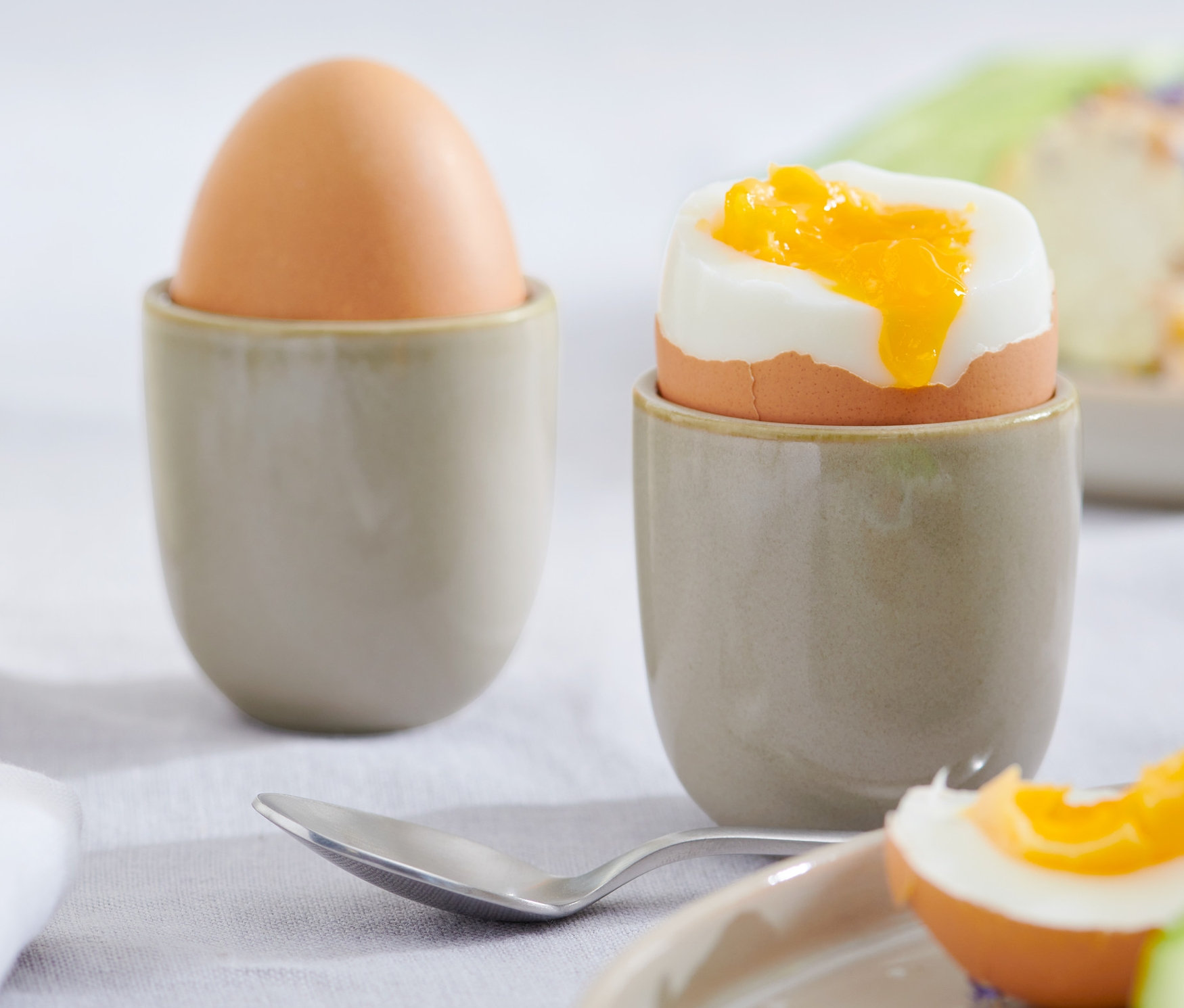Dwa jajka w jasnoszarych Kieliszkach do jajek, 4 sztuki. Jedno jajko jest całe, drugie jest ugotowane na miękko i przekrojone.