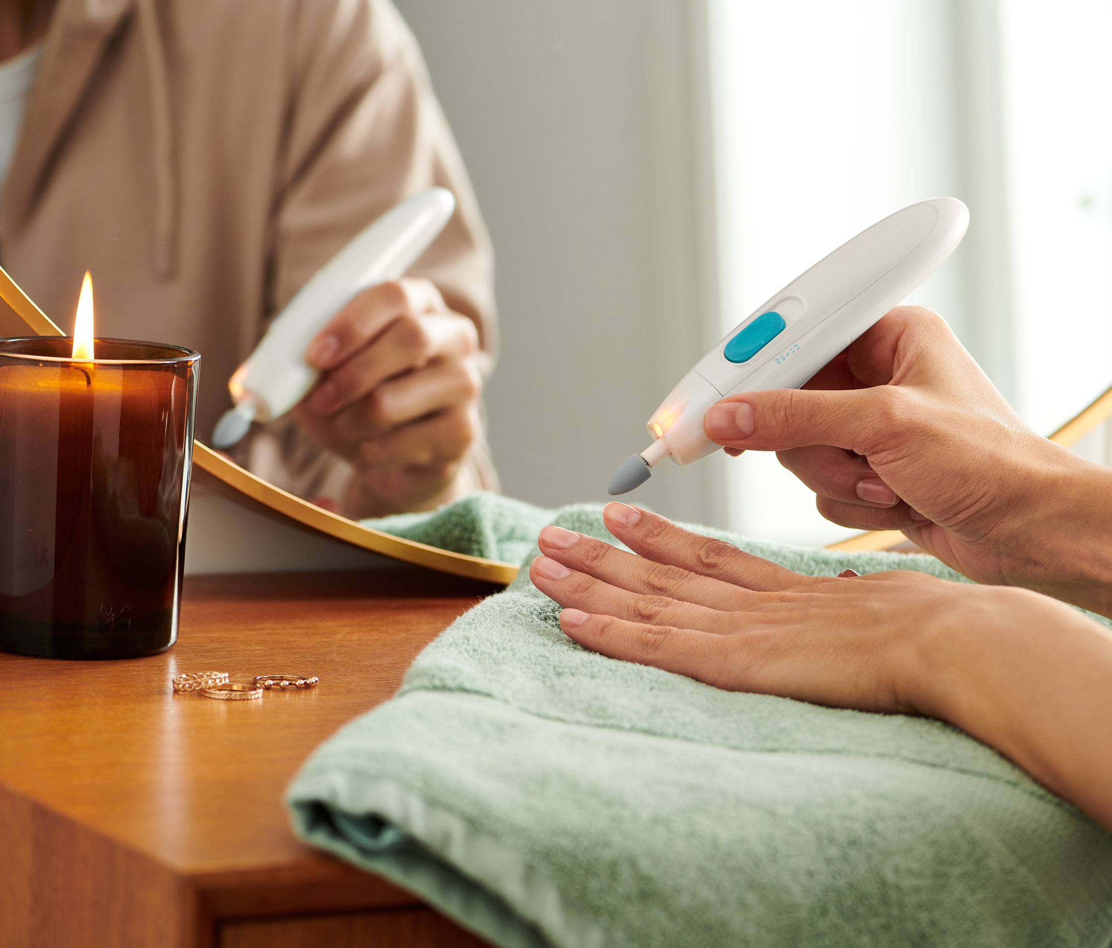 Kobieta używa urządzenia do manicure i pedicure na paznokcie. Na stole leży zielony ręcznik, świeca i pierścionki.