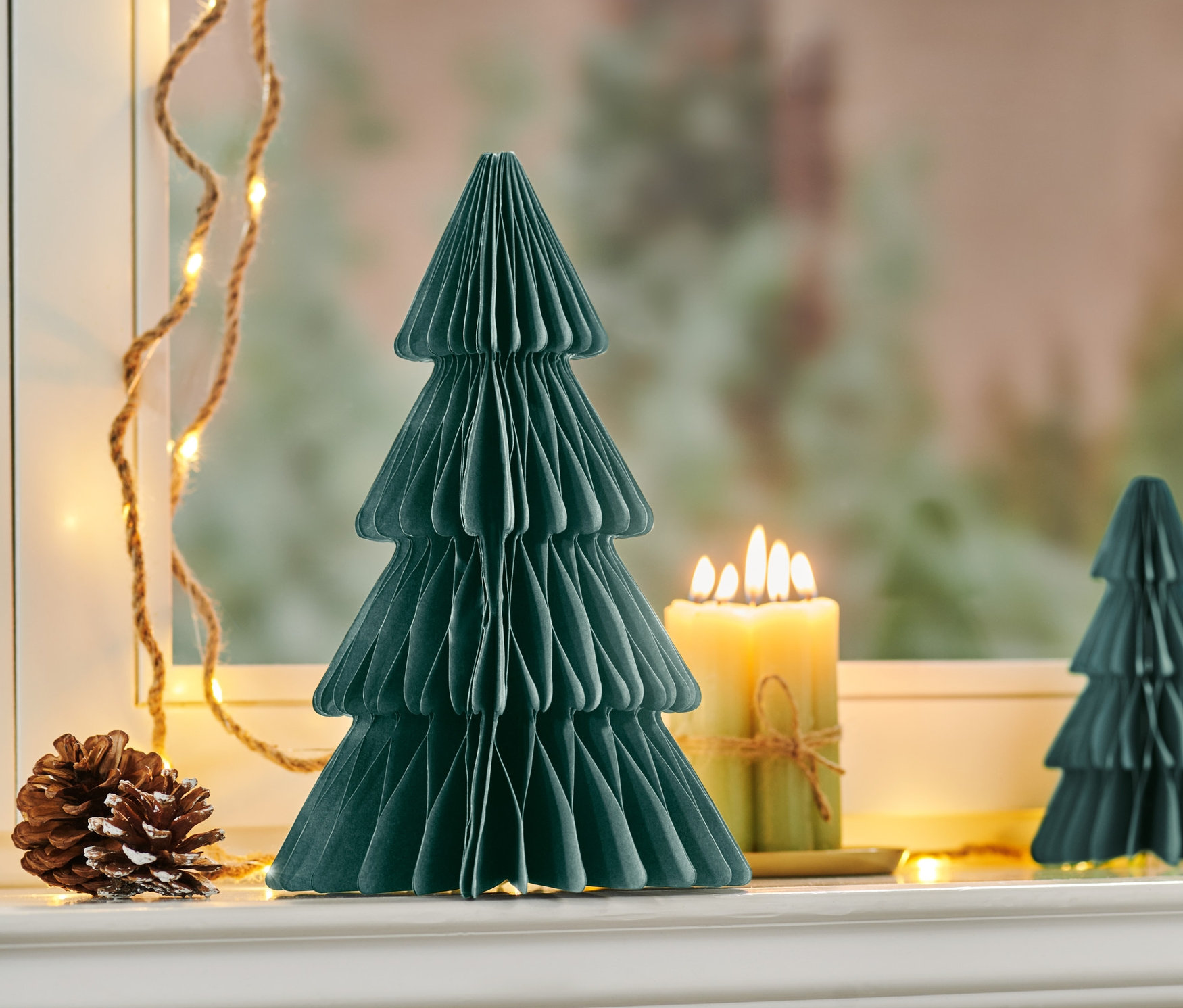 Średnia, ciemnozielona składana choinka z papieru stoi obok zapalonej świecy i szyszki na parapecie okna.