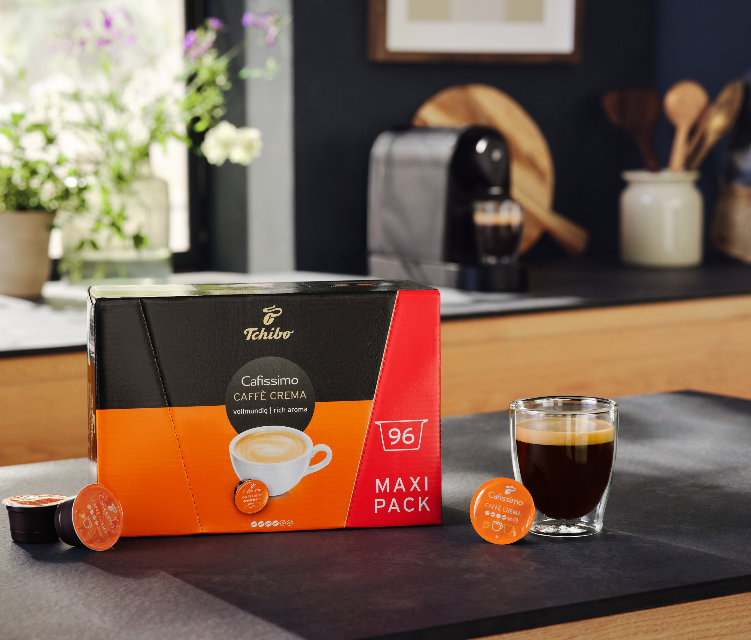 Opakowanie Cafissimo Caffè Crema Rich Aroma 96 kapsułek stoi obok szklanki kawy. W tle ekspres do kawy Cafissimo pure+, black.