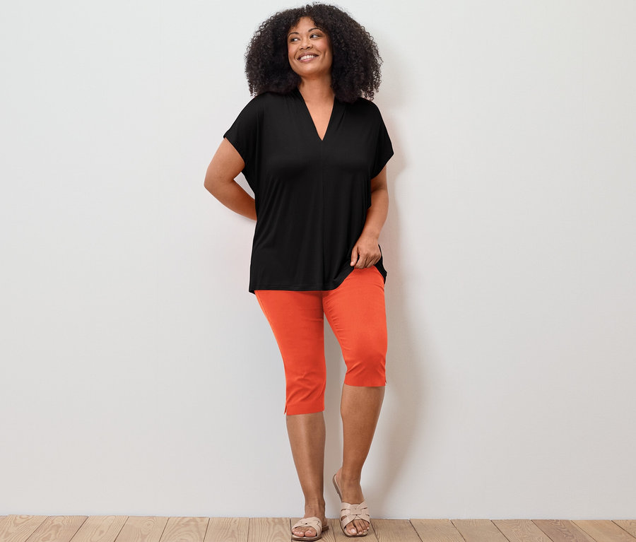 Kobieta pozuje w czarnej bluzce z dekoltem w serek i pomarańczowych spodniach stretchowych o długości 3/4.