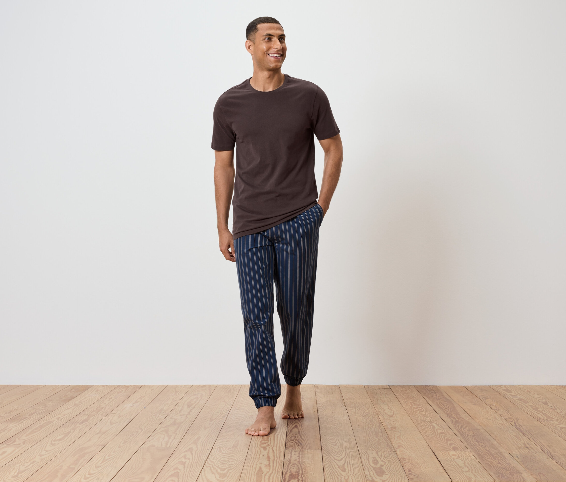 Mężczyzna idzie boso po drewnianej podłodze, ubrany w brązowy t-shirt i niebieskie spodnie od piżamy w paski.