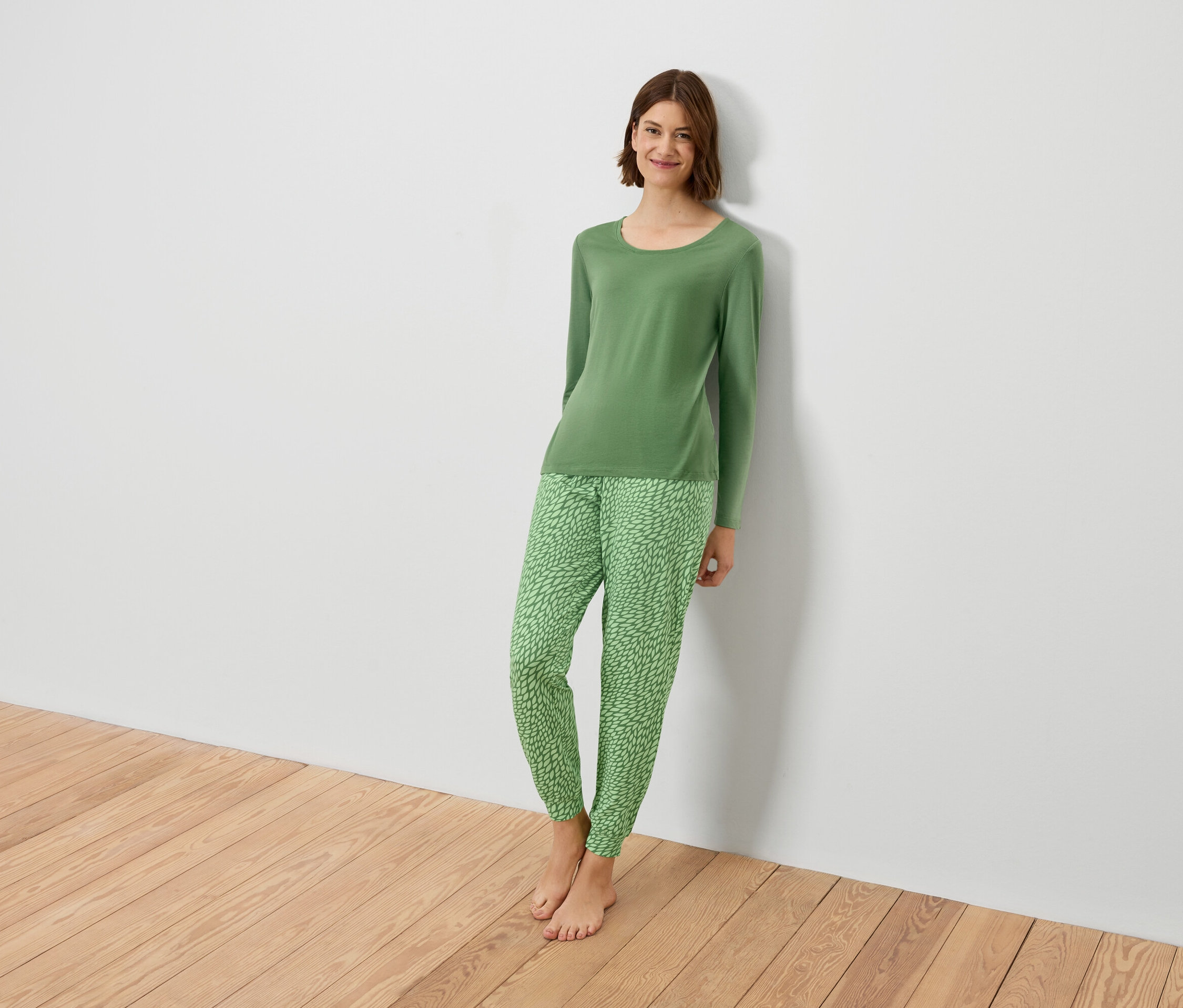 Kobieta w zielonej piżamie opiera się o białą ścianę.