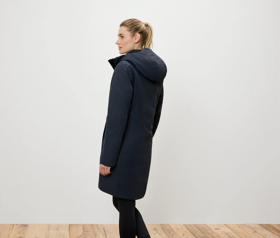 Widok z boku kobiety z blond włosami związanymi w kucyk, ubranej w długi, granatowy płaszcz z kapturem i czarne legginsy.