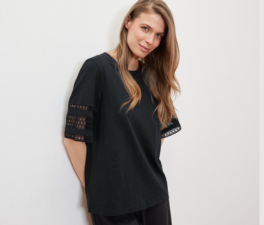 Kobieta oparta o ścianę ma na sobie czarną bluzkę z haftem.