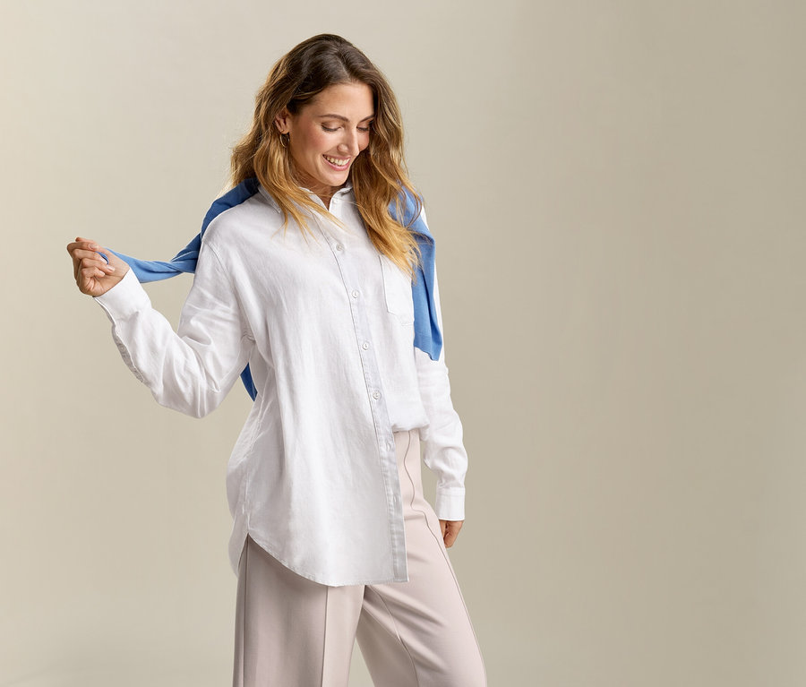 Uśmiechnięta kobieta z niebieskim swetrem na ramionach ma na sobie długą, białą lnianą bluzkę.