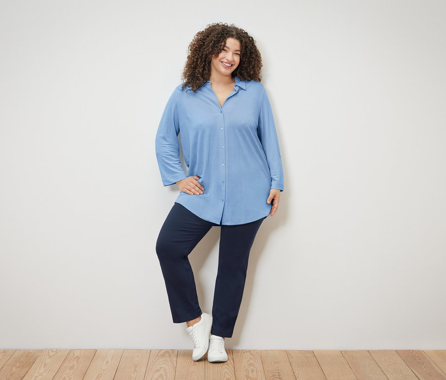 Kobieta z kręconymi włosami ma na sobie jasnoniebieską bluzkę, granatowe spodnie i białe buty.