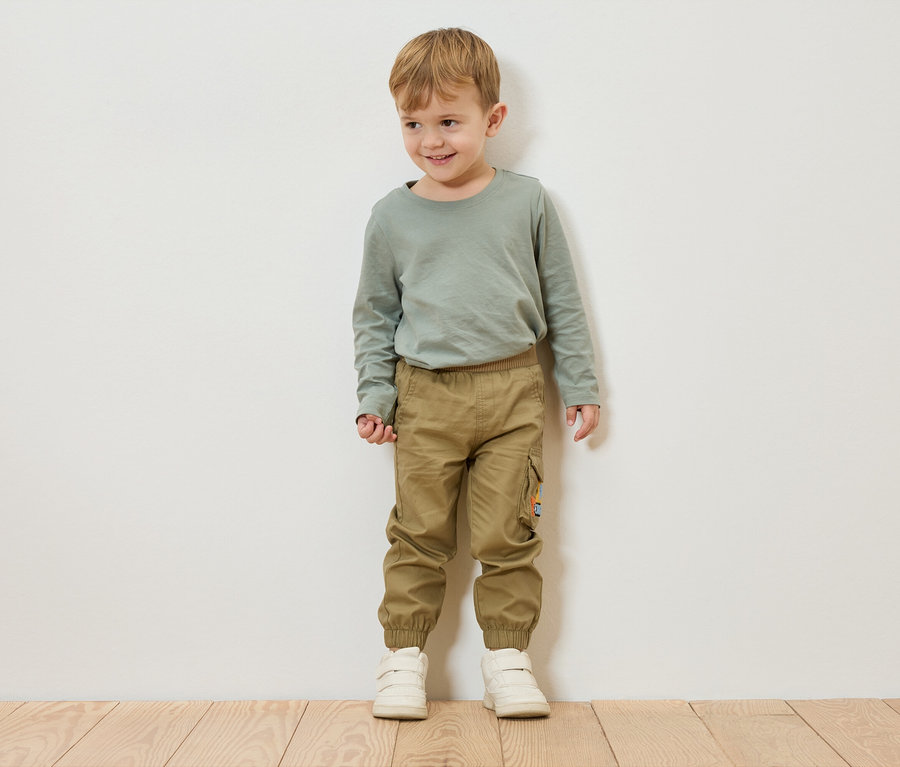 Chłopiec o blond włosach stoi na tle białej ściany, ubrany w zieloną koszulkę z długim rękawem, khaki spodnie i białe buty.