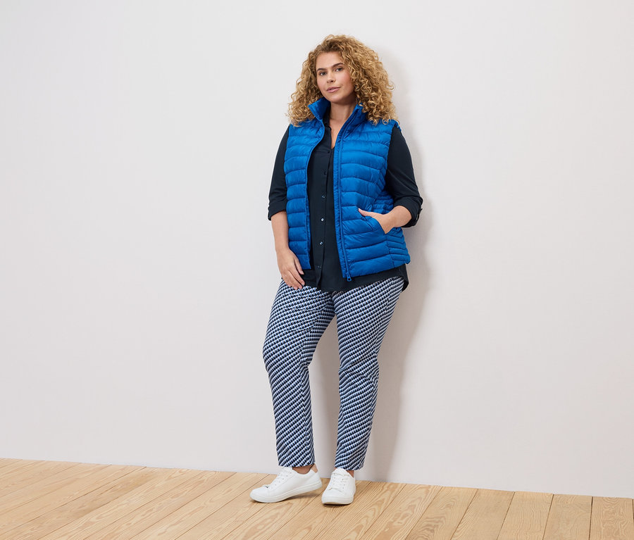 Kobieta o kręconych włosach w niebieskiej kamizelce, ciemnej koszuli i wzorzystych spodniach stoi przy białej ścianie.