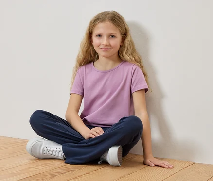 Dziewczyna z blond włosami siedzi ze skrzyżowanymi nogami na drewnianej podłodze, ubrana w liliowy T-shirt i niebieskie spodnie.