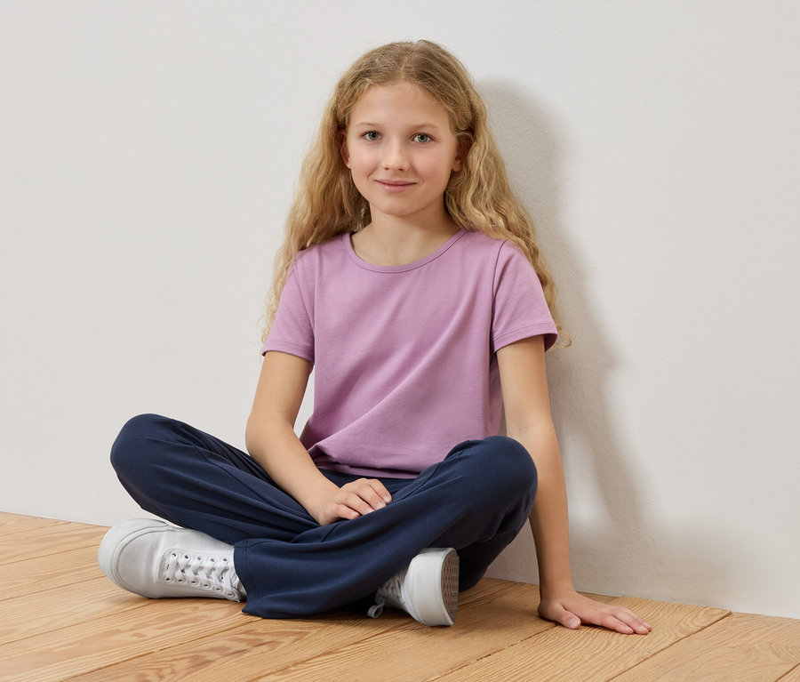 Dziewczyna z blond włosami siedzi ze skrzyżowanymi nogami na drewnianej podłodze, ubrana w liliowy T-shirt i niebieskie spodnie.