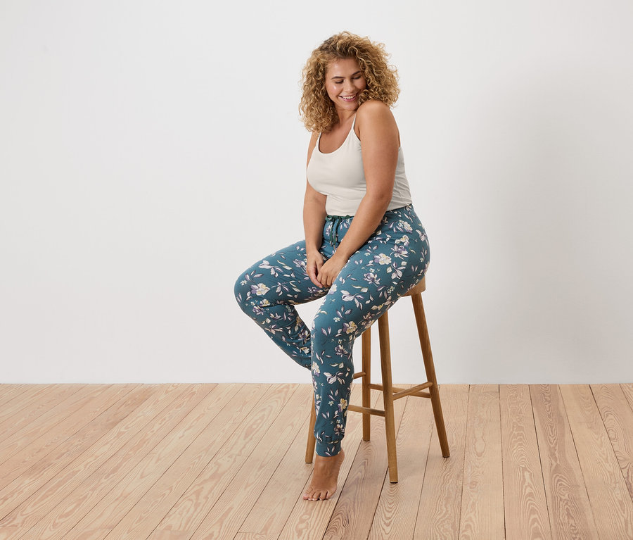 Kobieta z kręconymi włosami siedzi na stołku, ubrana w podkoszulek i kwieciste legginsy.