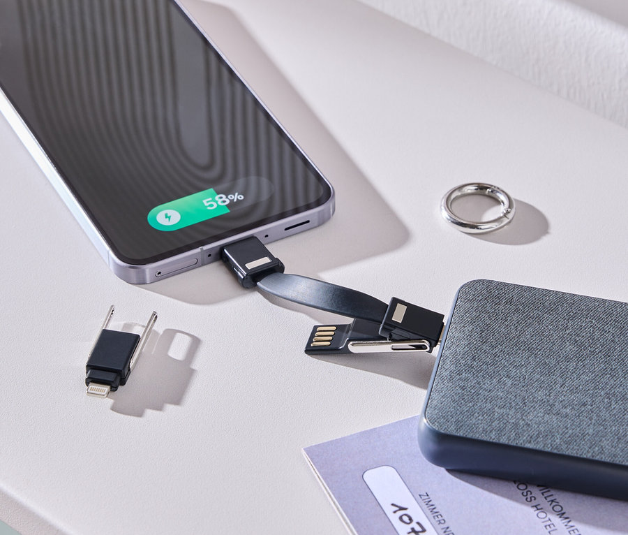 Smartfon ładowany za pomocą kabla i szarego akumulatora na białym stole. Obok leżą pierścionek i karta do pokoju.