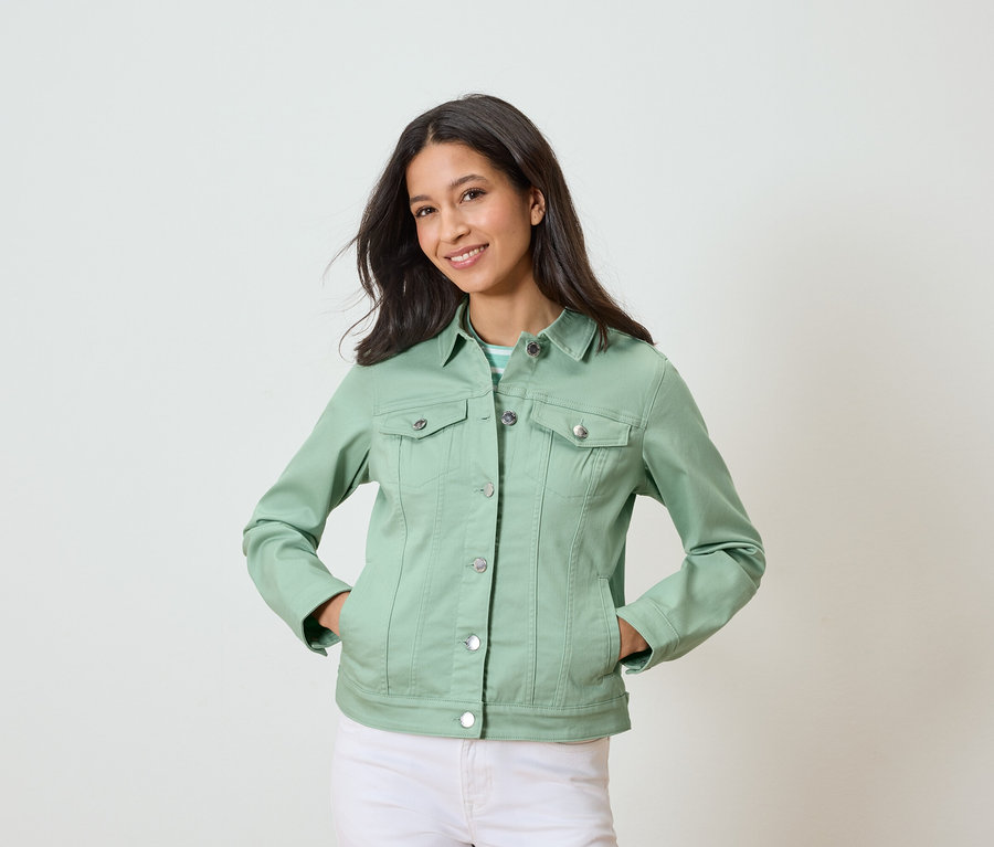 Kobieta pozuje z rękami w kieszeniach, ubrana w jasnozieloną kurtkę dżinsową i białe spodnie.