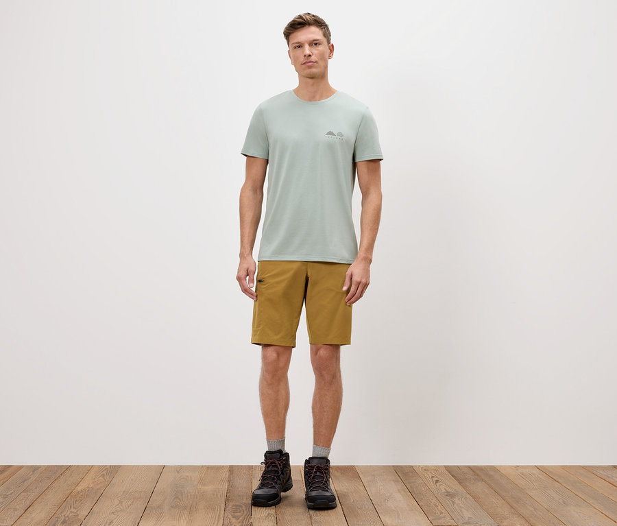 Mężczyzna stoi na tle białej ściany w zielonej koszulce, szortach i butach.