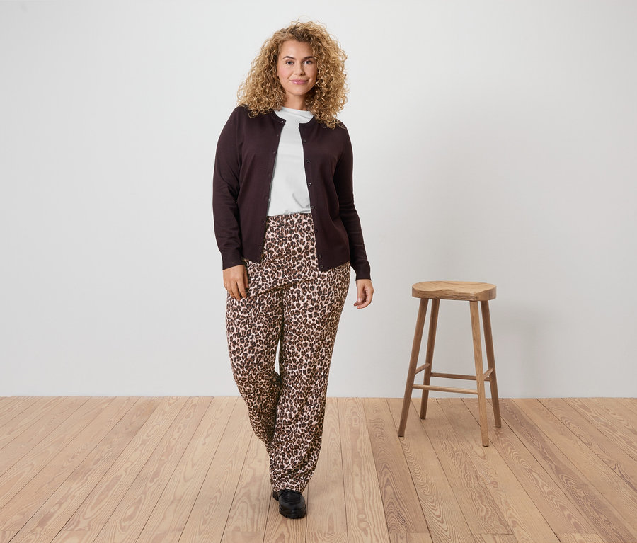 Kobieta o kręconych włosach ma na sobie brązowy kardigan, biały T-shirt i spodnie w panterkę.